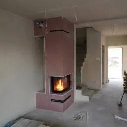 Wnętrze w trakcie wykańczania z zamontowanym kominkiem obudowanym płytami kartonowo-gipsowymi, widoczne przewody wentylacyjne i instalacyjne, na podłodze leżą płyty gipsowe.