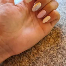 Dłoń z pomalowanymi paznokciami w odcieniach beżu, różu i srebra, z efektem brokatu i pęknięć, na tle marmurowego blatu.