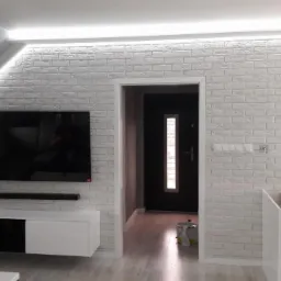 Salon z oświetleniem LED wokół sufitu i ściany z białej cegły dekoracyjnej, telewizor na ścianie, biała szafka RTV i nowoczesny żyrandol