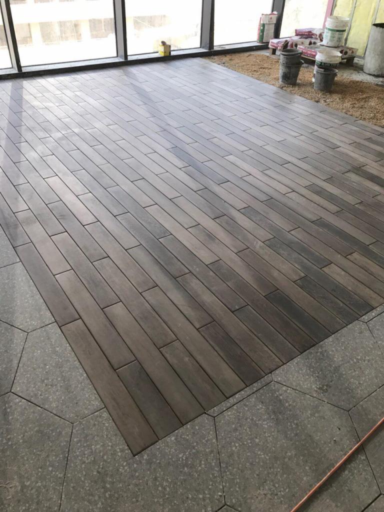 Układanie podłogi z płytek imitujących drewno w Ząbkach, z widocznym fragmentem posadzki z szarych, heksagonalnych płytek. W tle widoczne wiadra i worki z materiałami budowlanymi.