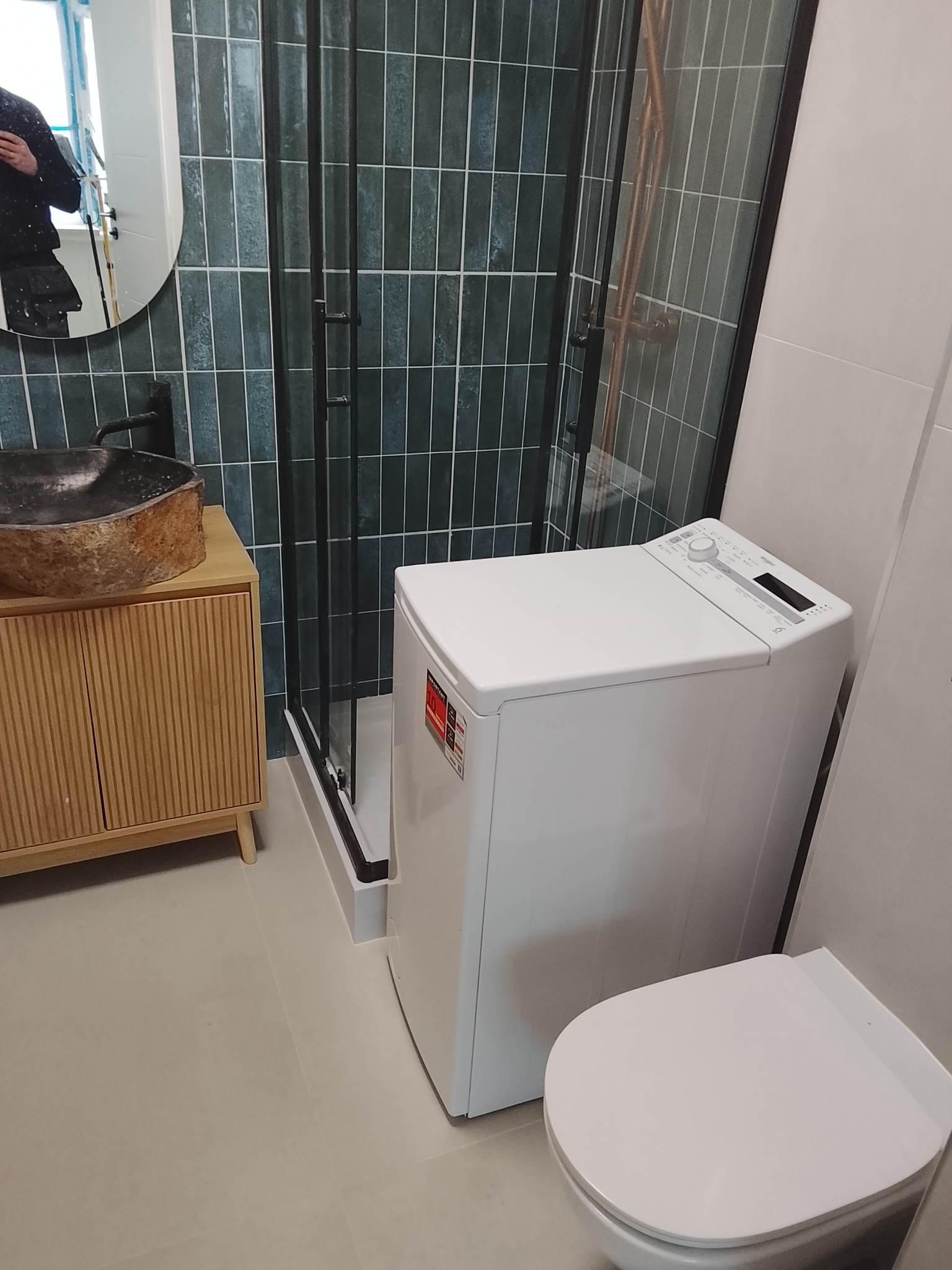 Nowoczesna łazienka z kabiną prysznicową, umywalką na szafce z lamelami i pralką. Ściany wyłożone podłużnymi, zielonymi płytkami. Widoczne odbicie osoby w lustrze.