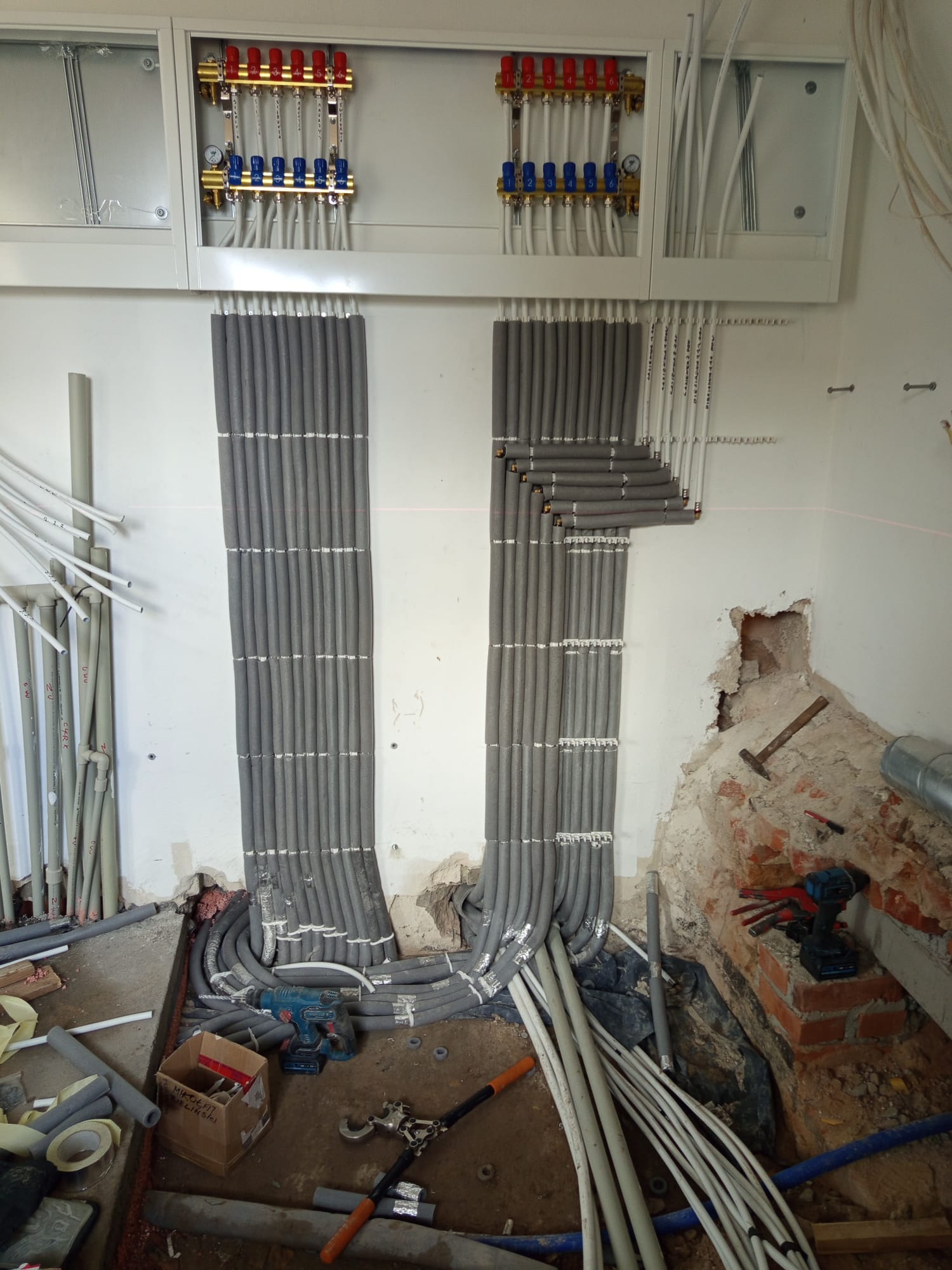 Rozdzielacz ogrzewania podłogowego z rurami w izolacji, widoczne narzędzia i elementy instalacji w trakcie montażu. Ściany w stanie surowym, widoczny laser krzyżowy.