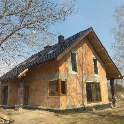 Niedokończony dom z cegły z nowym, czarnym dachem z dachówki, widoczne okna bez szyb i elementy więźby dachowej, na pierwszym planie paczki z materiałami budowlanymi, w tle drzewa bez liści...