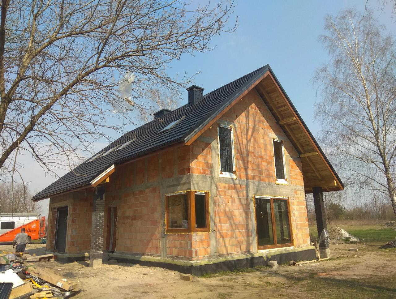 Niedokończony dom z cegły z nowym, czarnym dachem z dachówki, widoczne okna bez szyb i elementy więźby dachowej, na pierwszym planie paczki z materiałami budowlanymi, w tle drzewa bez liści...