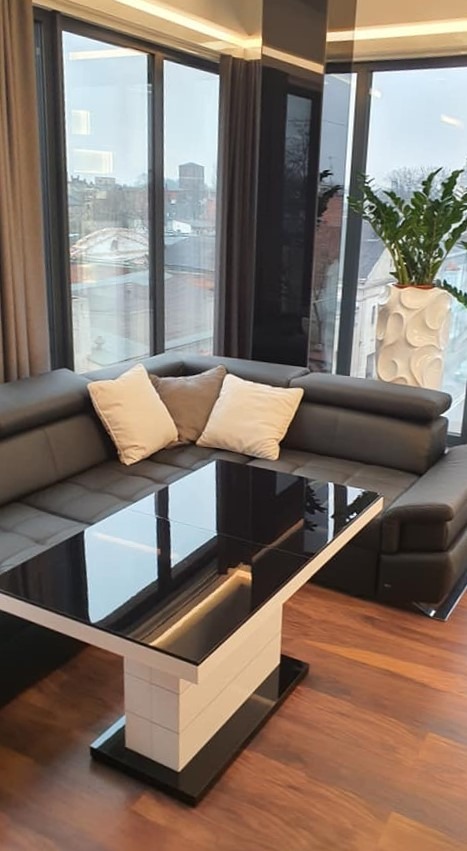 Nowoczesny salon z narożną sofą w odcieniach szarości, stolikiem kawowym z czarnym blatem i białymi elementami oraz dużymi oknami z widokiem na miasto i dekoracyjną donicą.