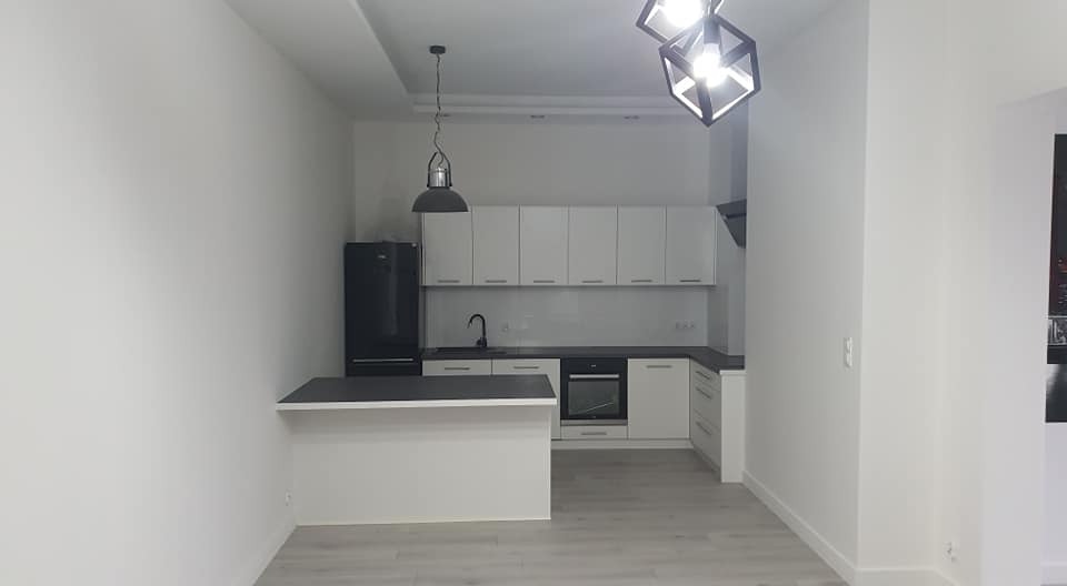 Nowoczesna, minimalistyczna kuchnia z białymi szafkami, czarnym blatem i lodówką, oświetlona wiszącymi lampami o geometrycznych kształtach.