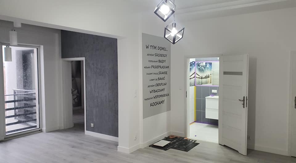 Jasne, przestronne wnętrze po remoncie z szarą ścianą akcentującą, ozdobnym napisem na ścianie, nowoczesnymi lampami i widokiem na łazienkę z motywem krajobrazowym.