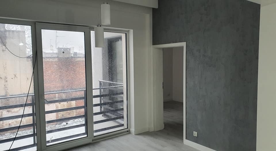 Nowoczesne wnętrze z szarą ścianą dekoracyjną, przesuwne okno balkonowe z widokiem na zimowy krajobraz miejski, minimalistyczna lampa wisząca i jasna podłoga.