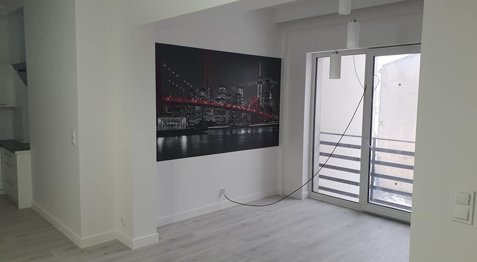 Jasne, nowocześnie wykończone wnętrze z szarymi panelami podłogowymi, białymi ścianami, dużym obrazem z nocnym widokiem miasta i balkonem z szarymi barierkami.