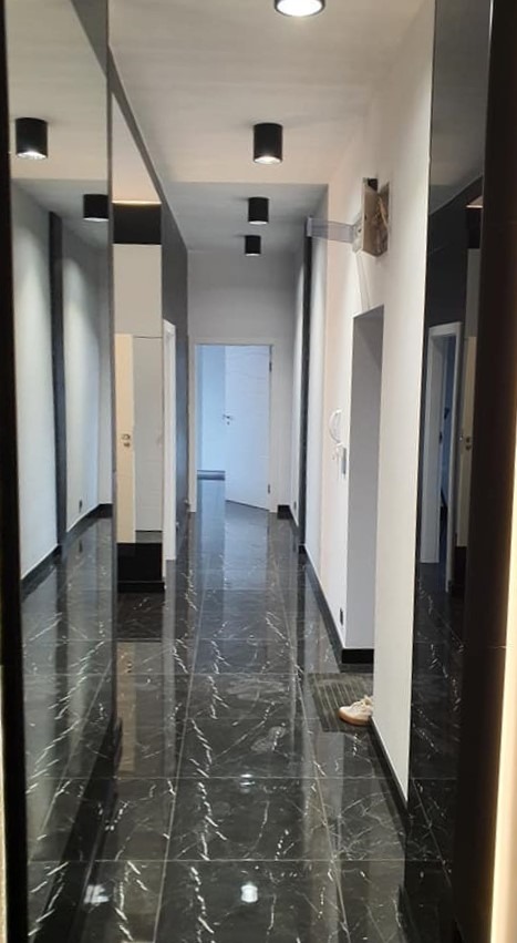 Długi korytarz z czarną marmurową podłogą, białymi ścianami i czarnymi akcentami, oświetlony cylindrycznymi lampami sufitowymi, z lustrami po bokach i pojedynczym butem przy ścianie.