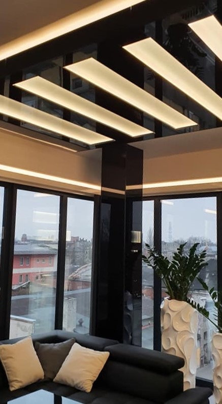 Nowoczesny salon z czarnym, podwieszanym sufitem z podświetleniem LED, narożną sofą, dużymi oknami z widokiem na miasto i dekoracyjnymi donicami z roślinami.