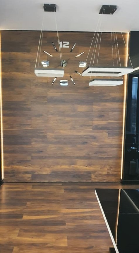 Nowoczesne wnętrze z drewnianą ścianą, podświetlanymi listwami LED, designerskim zegarem ściennym i wiszącymi lampami w kształcie kwadratów.