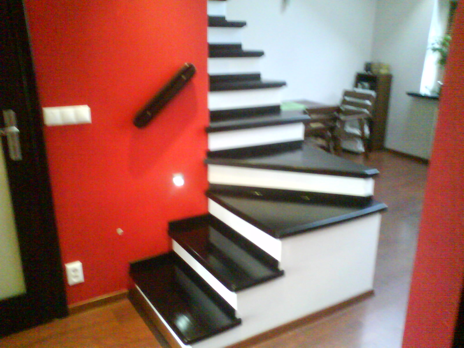 Nowoczesne schody z czarnymi stopniami i białymi podstopnicami, kontrastujące z czerwoną ścianą i ciemnobrązową poręczą, widoczne gniazdko elektryczne i włączniki światła.