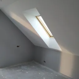 Szare wnętrze poddasza ze skośnym oknem dachowym, widoczne ślady po pracach wykończeniowych na podłodze i wystające przewody elektryczne.
