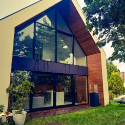 Nowoczesny dom z dużą, przeszkloną ścianą; widoczne ślady po deszczu na szybach, drewniana elewacja i zadbany trawnik.