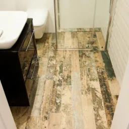 Ukończona łazienka z podłogą z płytek imitujących drewno, kabina prysznicowa ze szklanymi drzwiami i biała umywalka z czarną szafką.