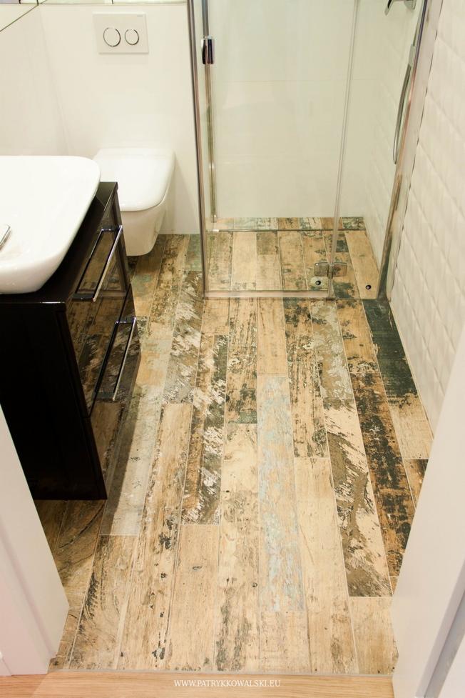 Ukończona łazienka z podłogą z płytek imitujących drewno, kabina prysznicowa ze szklanymi drzwiami i biała umywalka z czarną szafką.