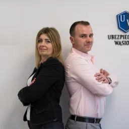 Dwoje agentów ubezpieczeniowych, kobieta w czarnej marynarce i mężczyzna w różowej koszuli, stoją plecami do siebie na tle białej ściany z logo firmy 'Ubezpieczenia Wasiołek'.