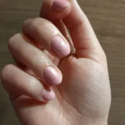 Dłoń z delikatnym, różowym manicure z subtelnym, błyszczącym pyłkiem, na tle drewnianej powierzchni.