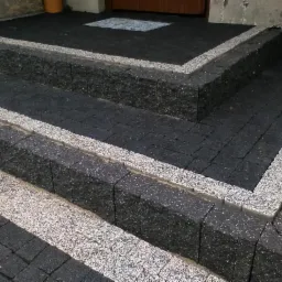 Schody wejściowe wyłożone czarną kostką brukową z obramowaniem z jasnej kostki, prowadzące do drzwi wejściowych.