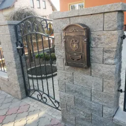 Otwarta, ozdobna, metalowa furtka z zamkiem, przylegająca do murowanego słupka z szarej kostki, na którym zamontowano stylizowaną skrzynkę na listy w kolorze starego złota.