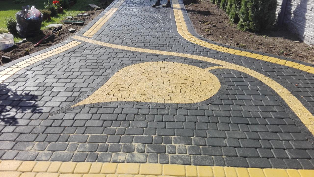 Ścieżka z ciemnoszarej kostki brukowej z żółtymi akcentami w kształcie liścia i obramowania, widoczne buty osoby w oddali.