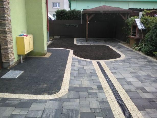 Ułożona nawierzchnia z kostki brukowej w odcieniach szarości, z akcentami w kolorze piaskowym, tworząca wzór z odpływem liniowym. W tle widoczna altana ogrodowa i fragment domu z żółtą szafką...