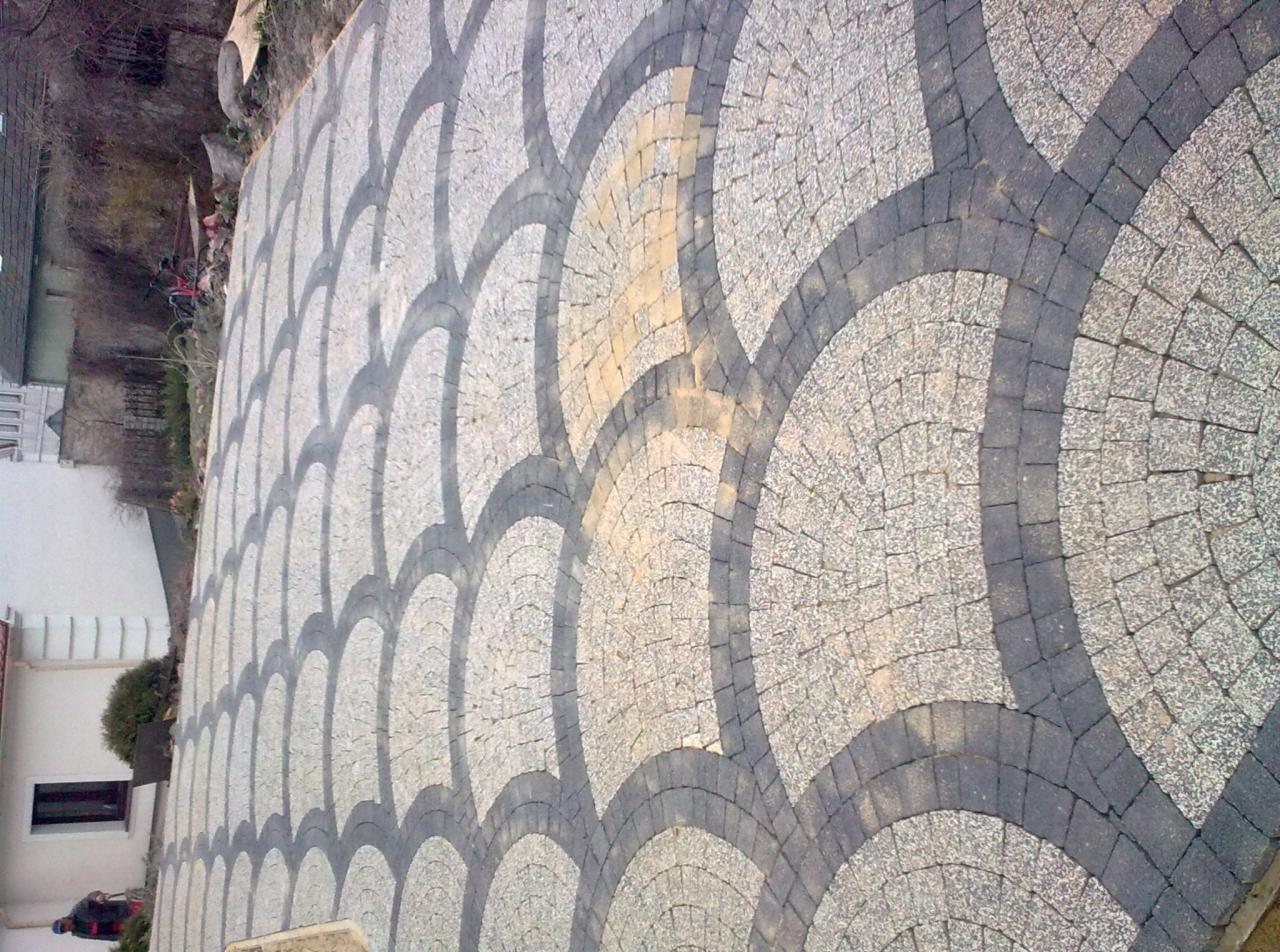 Ułożona kostka granitowa w kształcie wachlarza, z ciemnoszarymi obramowaniami, przed budynkiem z białymi ścianami i krzewem.