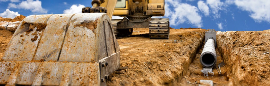 Żółta koparka z łyżką w wykopie obok ułożonej rury kanalizacyjnej z betonu, widok z dołu na tle błękitnego nieba z chmurami.