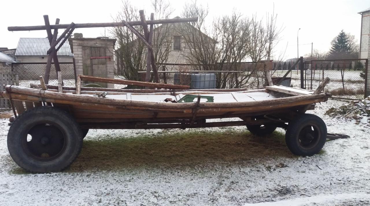 Stary, drewniany wóz drabiniasty na tle zimowego krajobrazu. Wóz z dużymi kołami, częściowo pokryty śniegiem, stoi na trawie. Widoczny budynek w tle.