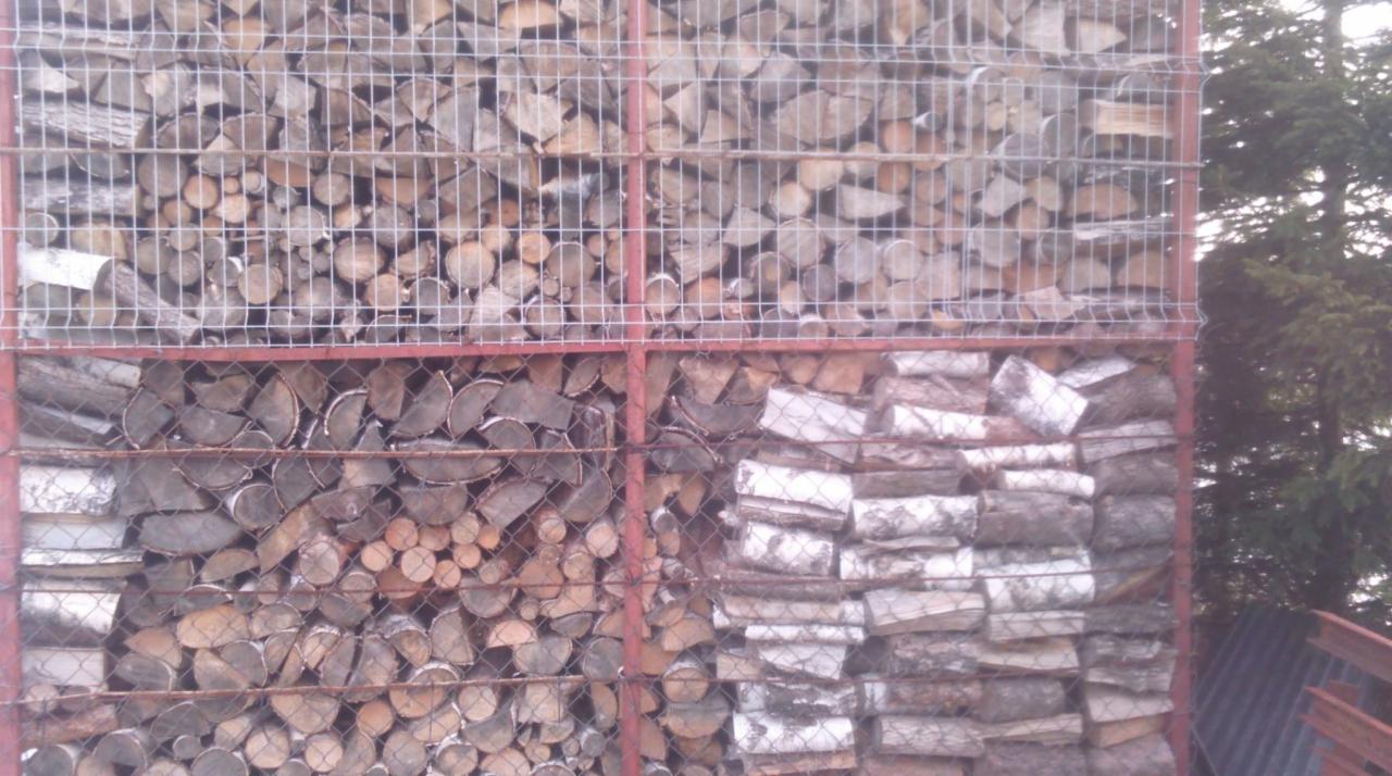 Metalowa konstrukcja wypełniona pociętymi kawałkami drewna opałowego, widoczne różne gatunki i kształty, tło z zielenią drzew.