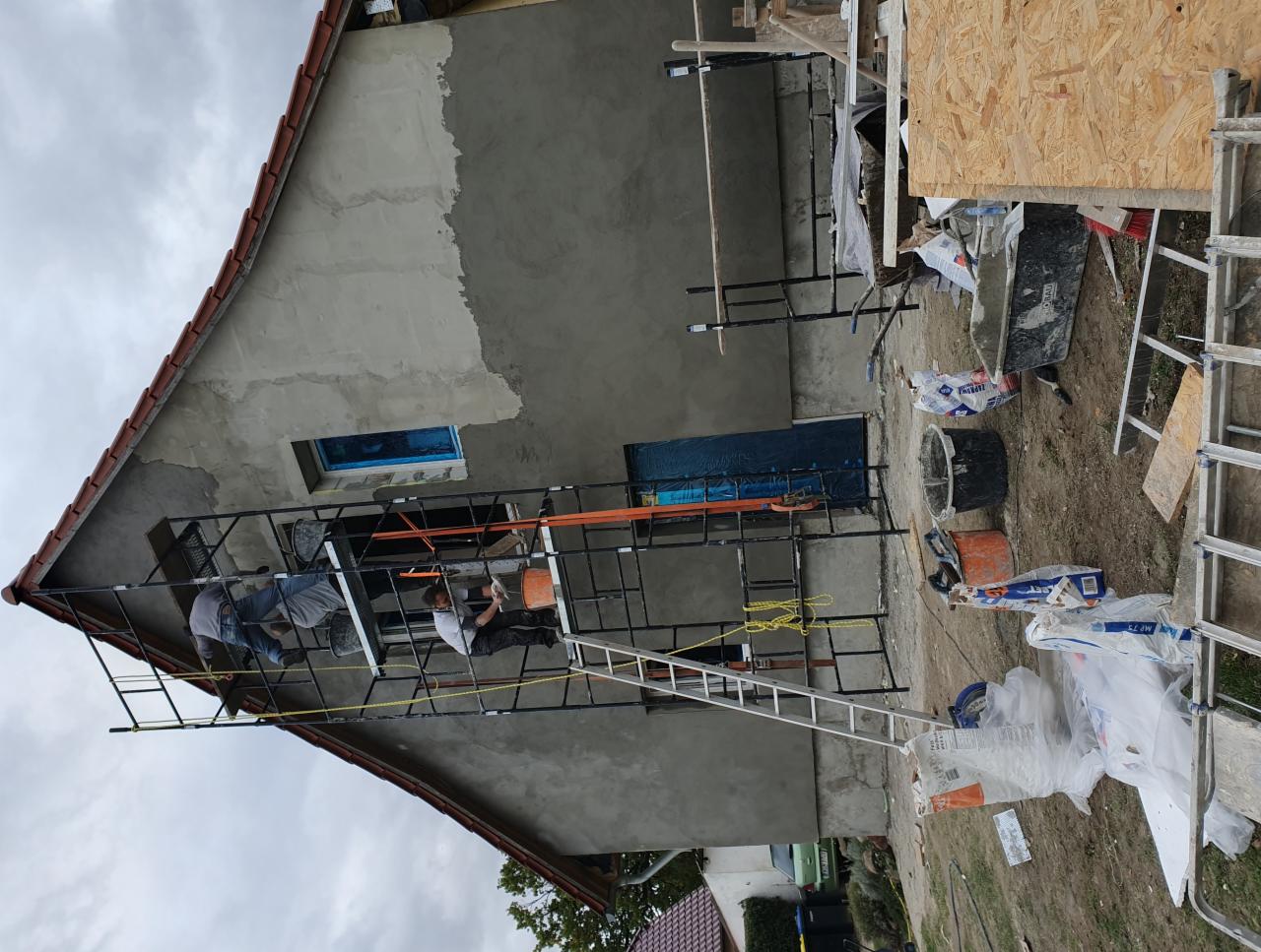 Dwóch pracowników na rusztowaniu podczas nakładania szarego tynku na elewację domu z czerwoną dachówką, widoczne okno zabezpieczone folią, materiały budowlane i drabina na ziemi.