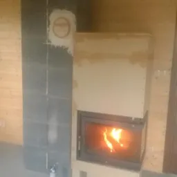 Wnętrze z kominkiem w trakcie montażu, widoczny ogień w palenisku. Obok kominka stoi butla z pianką montażową. Ściany wyłożone drewnianą boazerią.