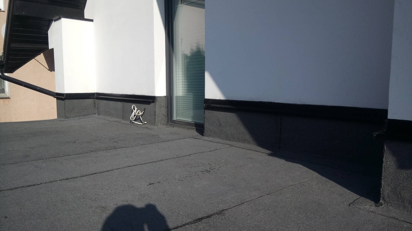 Widok na płaski dach pokryty papą, z widoczną ścianą budynku z oknem i zwisającym przewodem elektrycznym.