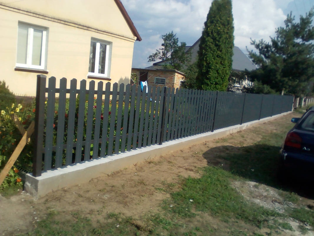 Nowo postawione, szare, drewniane ogrodzenie panelowe z betonową podmurówką, oddzielające posesję z domem i ogrodem od drogi.