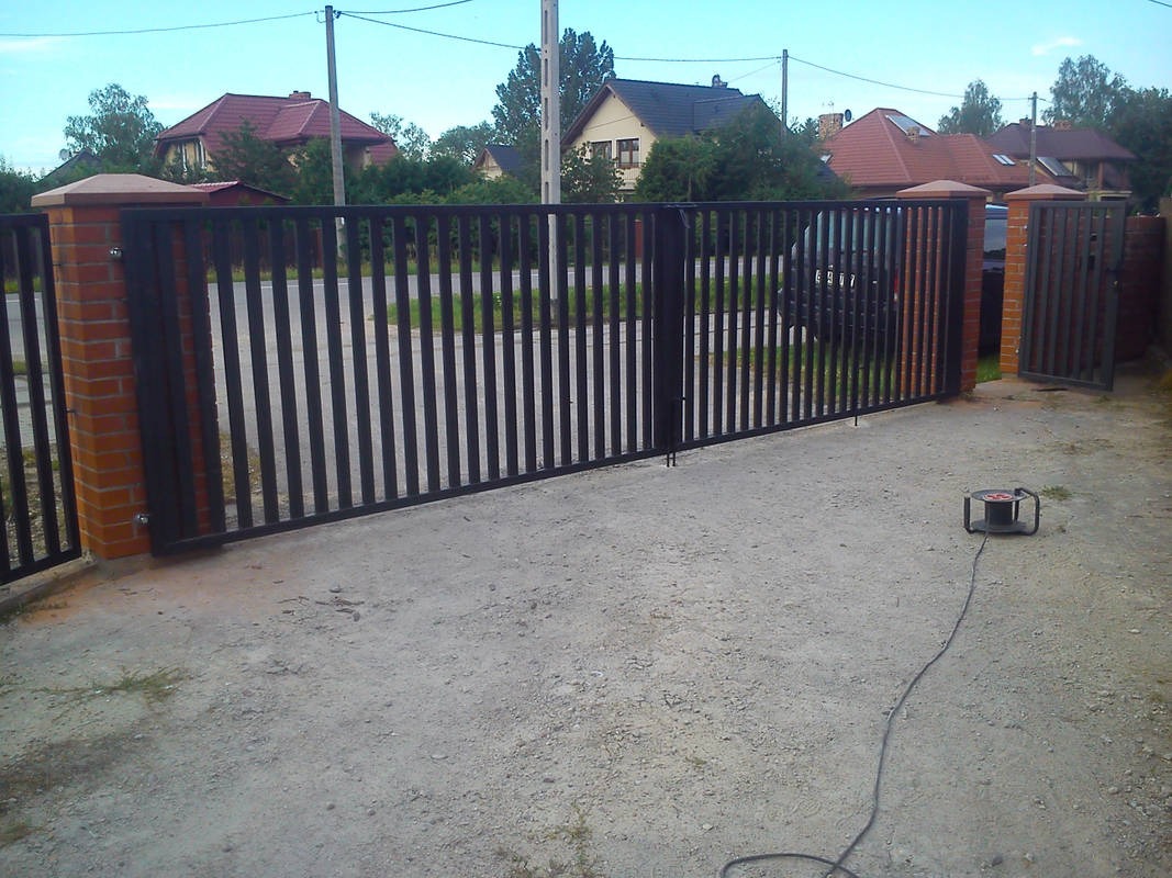 Nowoczesna, metalowa brama wjazdowa z pionowymi szczeblami, osadzona na ceglanych słupkach, widoczny zwinięty przedłużacz na żwirowym podłożu, w tle domy jednorodzinne.