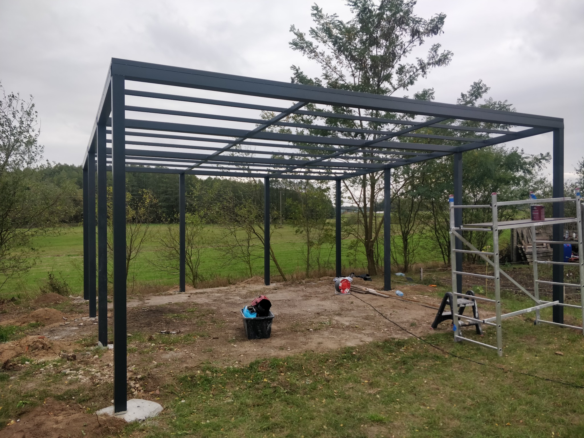 Stalowa konstrukcja pergoli w trakcie montażu na zewnątrz, widoczne narzędzia i rusztowanie. Szara, metalowa rama na tle zieleni drzew i pochmurnego nieba.