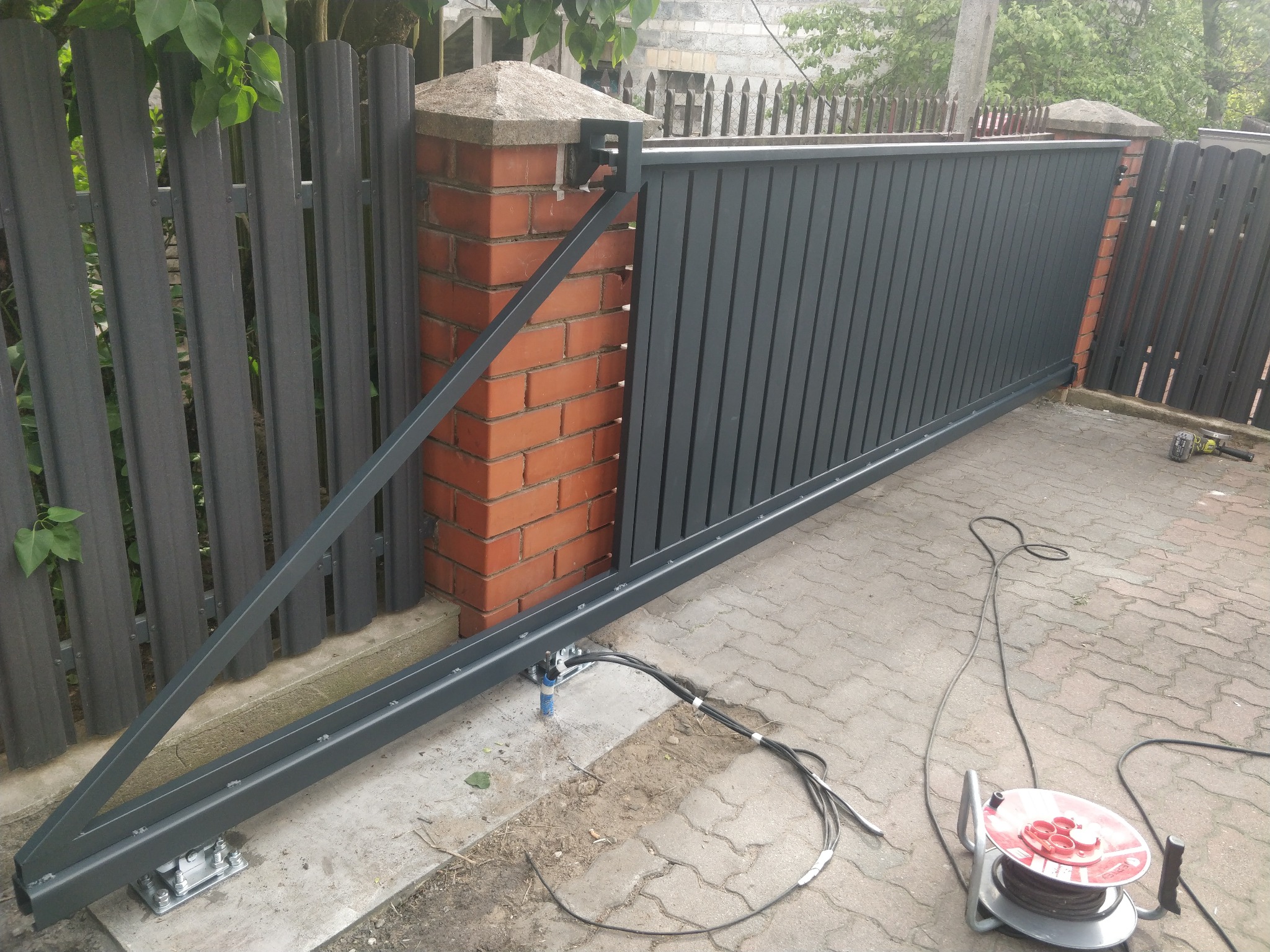 Nowoczesna, przesuwna brama wjazdowa z szarych paneli pionowych, zamontowana na ceglanym słupku. Widoczne okablowanie i narzędzia montażowe na kostce brukowej.