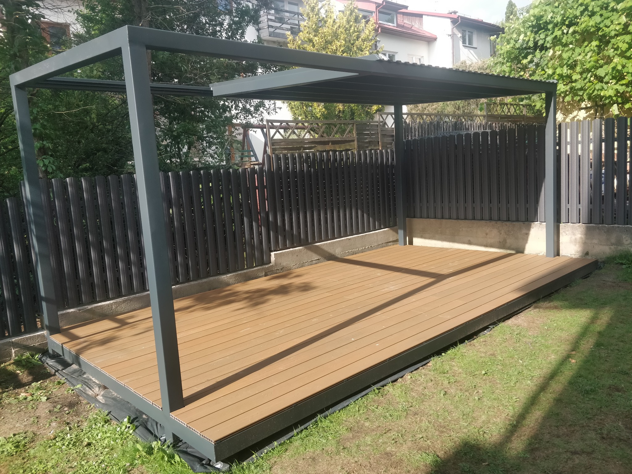 Nowoczesna pergola metalowa z drewnianym podestem na trawniku, szara konstrukcja, widok na ogrodzenie panelowe i budynek w tle, słoneczny dzień.
