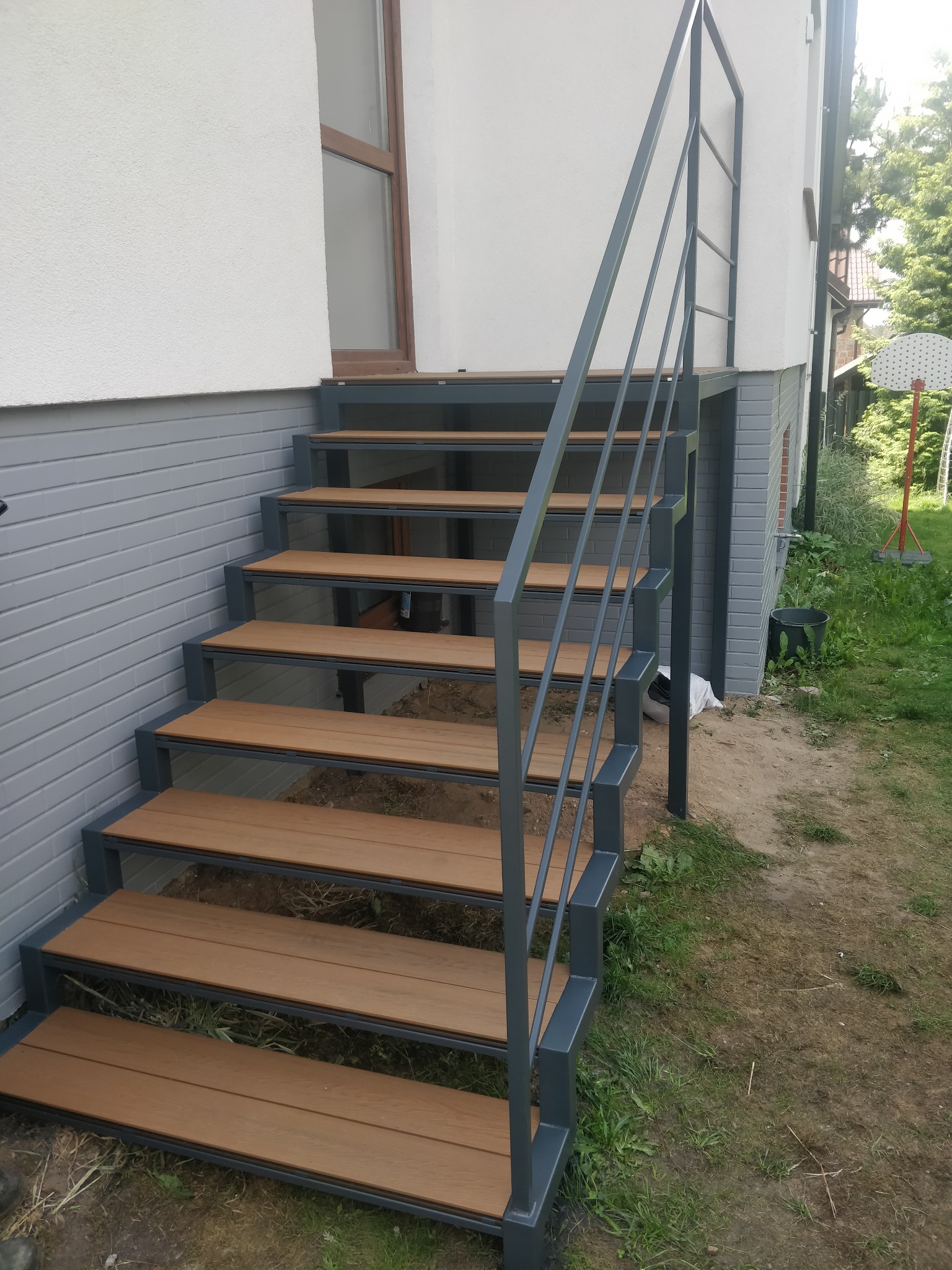 Zewnętrzne schody z metalową konstrukcją i drewnopodobnymi stopniami prowadzące do wejścia do domu. Szara kolorystyka konstrukcji i naturalne odcienie drewna.