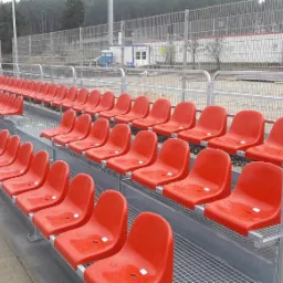 Nowo wybudowane, modułowe trybuny stadionowe z czerwonymi plastikowymi siedzeniami i metalową konstrukcją kratową, widok na ogrodzenie i budynek zaplecza w tle.