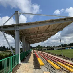 Zadaszona trybuna stadionu z konstrukcją stalową, widok z perspektywy chodnika, z zielonym ogrodzeniem i częściowo ułożonymi siedzeniami w kolorach czerwonym i żółtym.