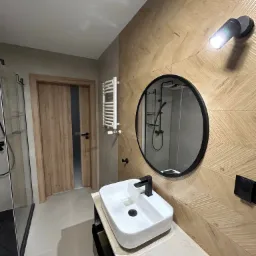 Nowoczesna łazienka z drewnopodobną ścianą w jodełkę, okrągłe lustro z czarną ramą, biała umywalka nablatowa z czarną armaturą, szklana kabina prysznicowa i grzejnik drabinkowy.