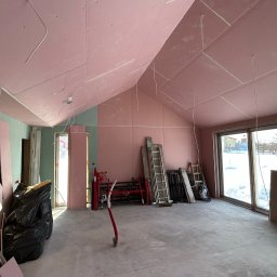 Jakubiec Adam - Wnętrze w trakcie prac wykończeniowych: ściany i sufit pokryte różowymi płytami kartonowo-gipsowymi, widoczne kable, drabiny i narzędzia, duże okno z widokiem na zaśnieżony ogród.