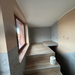 Jakubiec Adam - Wnętrze po remoncie: schody z drewnopodobnych paneli prowadzą na podest, ściany częściowo pomalowane na beżowo, częściowo z szarą strukturą, okno z brązową ramą.