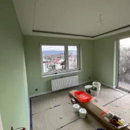 Świeżo pomalowane na pistacjowo ściany pokoju z widokiem na góry, zabezpieczona podłoga, pędzle i kuweta malarska na kartonie.