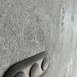 Szara, chropowata ściana z fakturą, z widocznym fragmentem czarnego przycisku spłuczki do WC i drobnymi, żółtymi przebarwieniami w jednym miejscu.