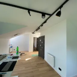 Wnętrze pokoju z drewnianą podłogą, świeżo pomalowanymi białymi ścianami i zamontowanym systemem szynoprzewodów z reflektorami. Widoczny fragment biurka z komputerem, grzejnik i drzwi wewnętrzne...