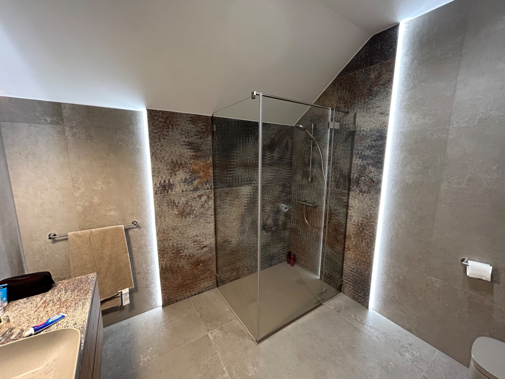 Nowoczesna łazienka z kabiną prysznicową typu walk-in, szare płytki imitujące beton, oświetlenie LED w szczelinach między płytkami, oraz widoczne akcesoria łazienkowe.