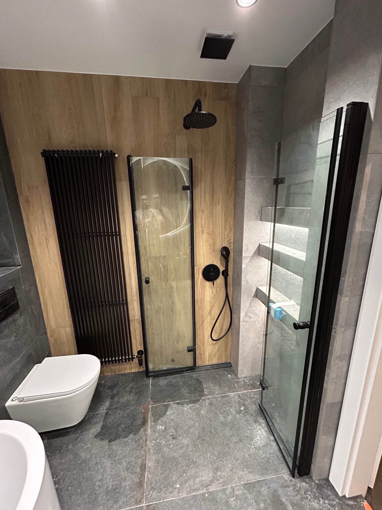 Nowoczesna łazienka z kabiną prysznicową typu walk-in z czarnymi profilami, grzejnikiem drabinkowym w kolorze czarnym, toaletą podwieszaną i drewnopodobną okładziną ścienną.